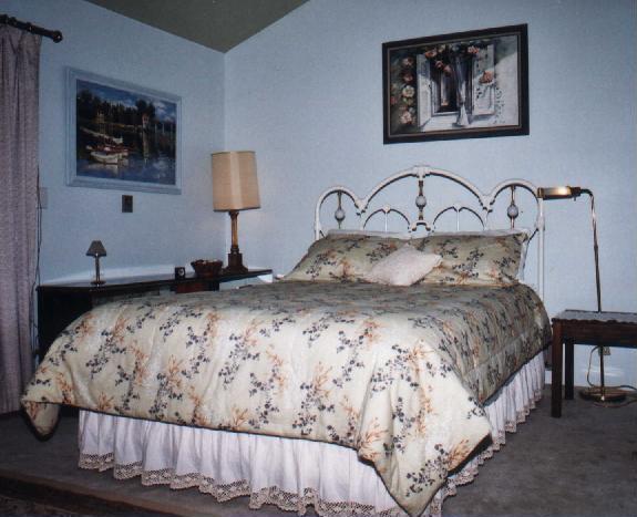Picture of the master bedroom, includes queen size bed with white iron victorian headboard, a picture on the wall of a boat on the water and a picture of flowers around a shuttered window